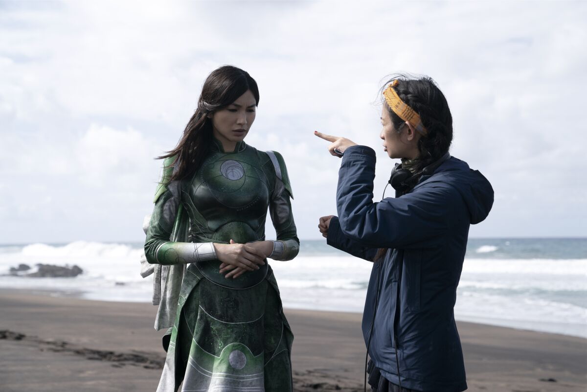 A woman in a blue jacket, right, talks to a woman in a green superhero suit on a beach.