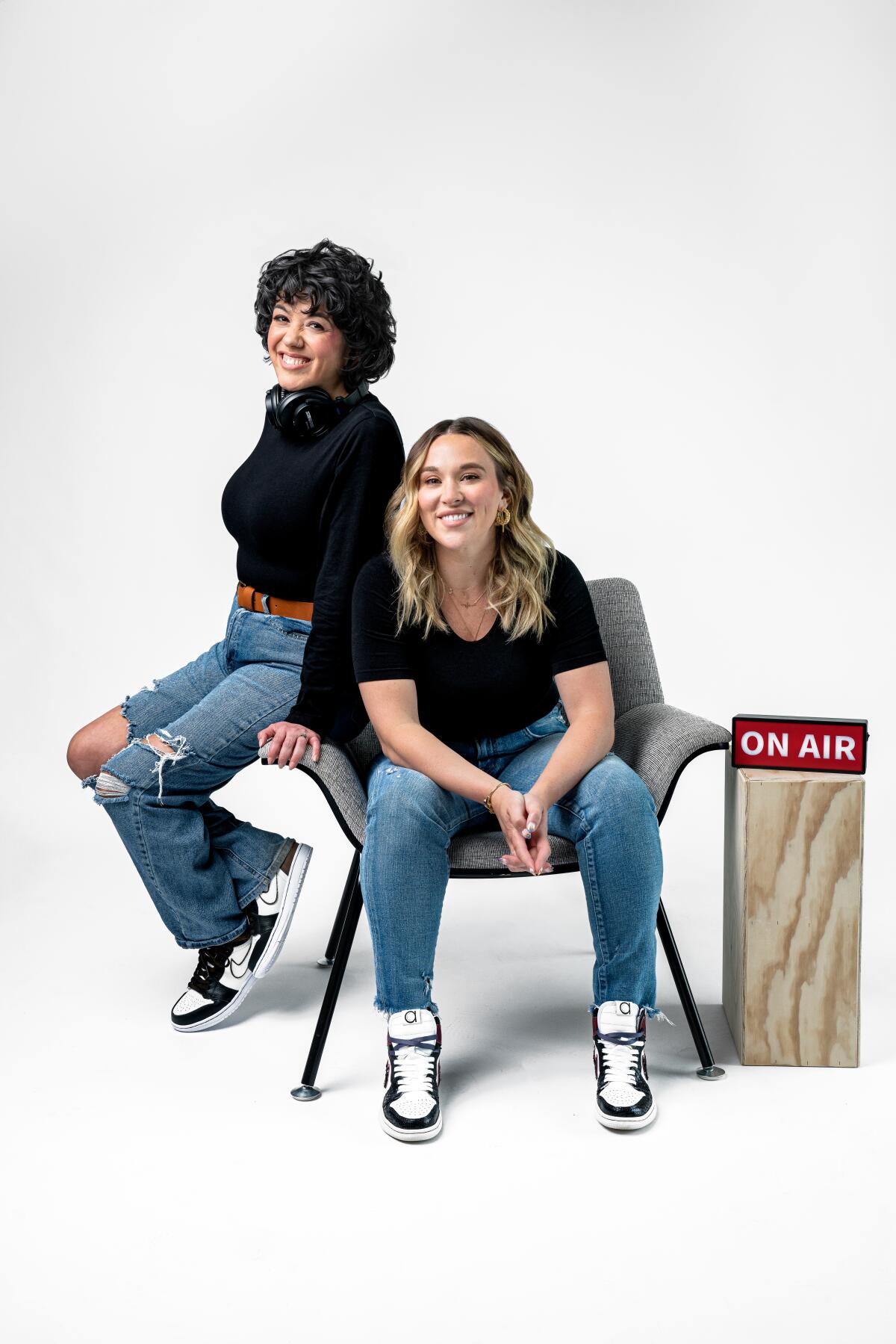 "Crime Junkie" hosts Brit Prawat, left, and Ashley Flowers sit in and on a chair by an "on air" sign.