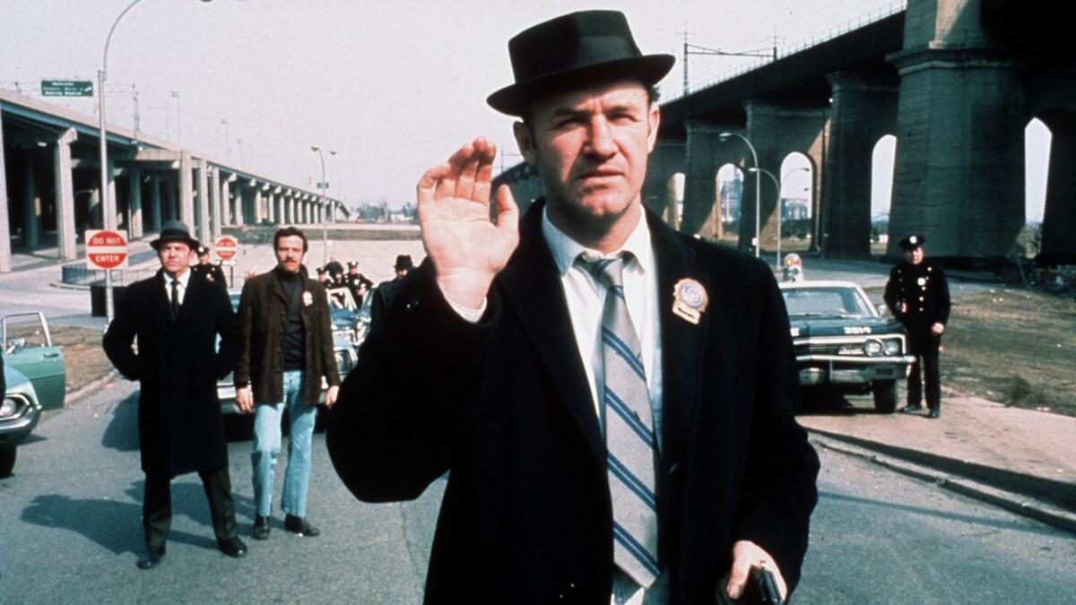 Gene Hackman holds up his right hand while wearing a hat and badge.