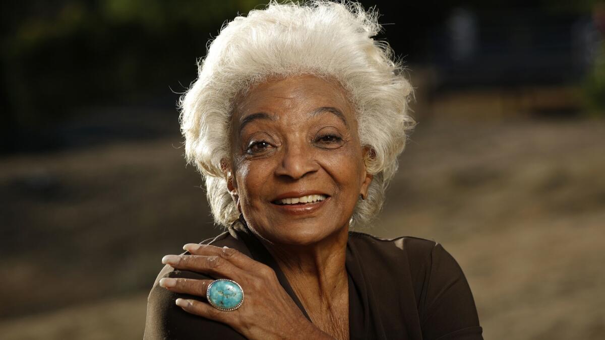  Nichelle Nichols smiles with one hand on her shoulder.