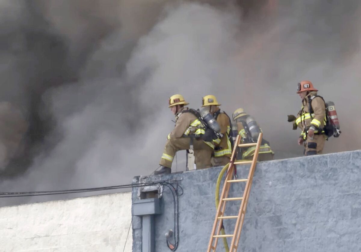 Los Angeles city firefighters work on the roof of a building while smoke billows behind them.