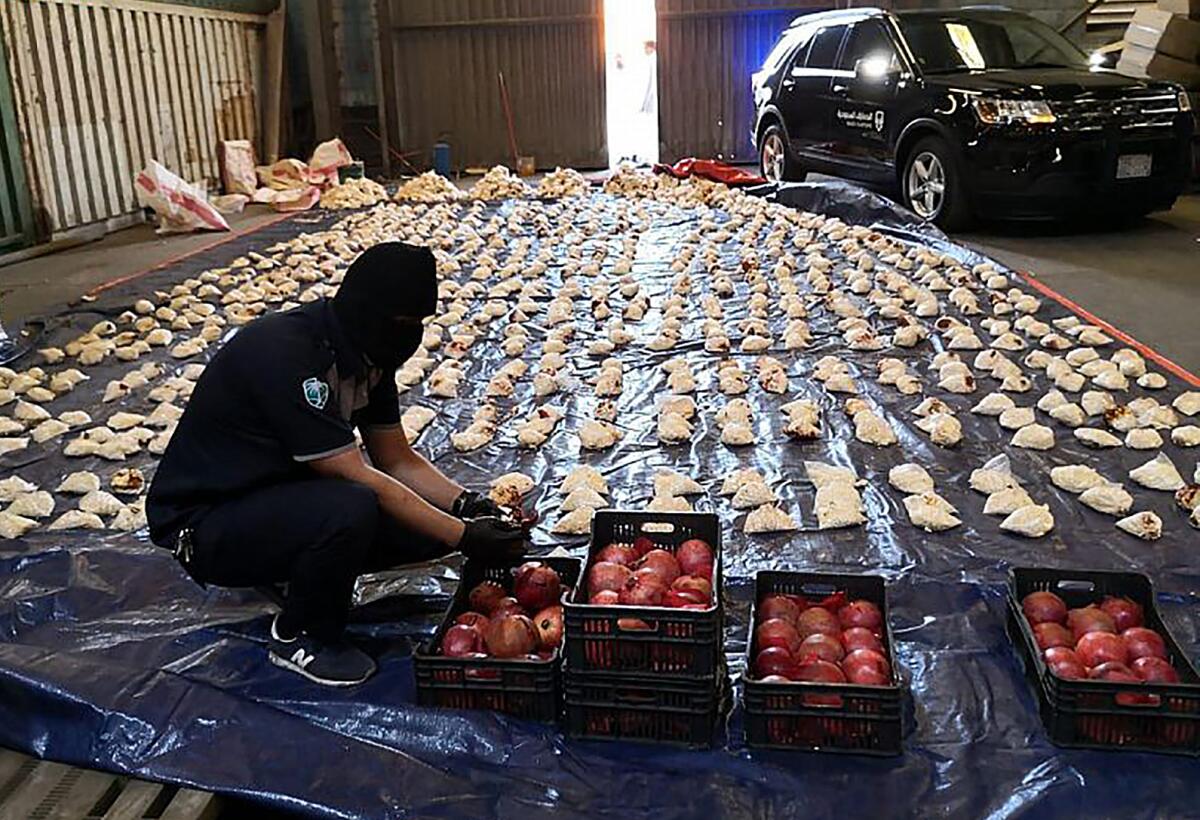 A person in dark uniform and mask examines boxes of pomegranates near multiple white bags laid out to his left