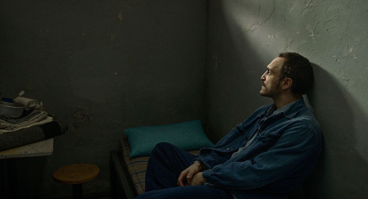A man sits on a cot, leaning against the wall, in a dark room 