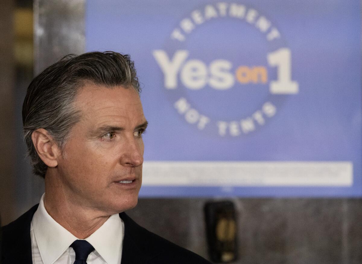 A man in dark suit and tie looks to the side. Behind him is a blue sign that reads Yes on 1