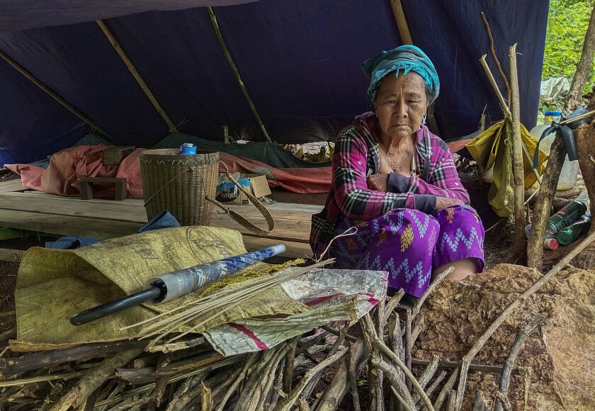 A woman in a blue head wrap and red-and-black plaid top sits near a pile of branches in a makeshift tent