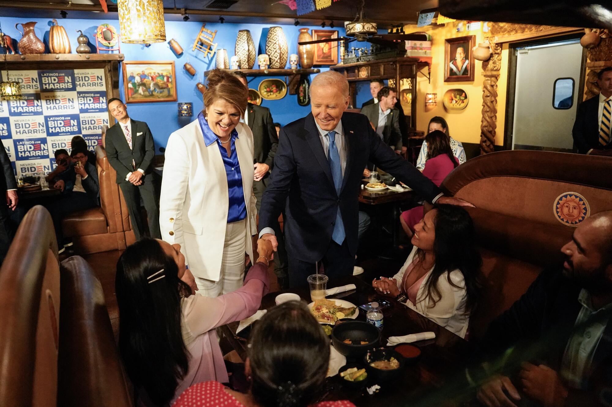 A woman in a white suit and blue blouse stands next to a man in dark suit and blue tie shaking the hand of restaurant patron