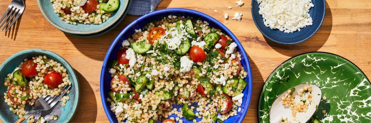 Overhead view of salad of tomatoes, cucumbers and Israeli couscous with feta and lots of lemon in four bowls on a tabletop.