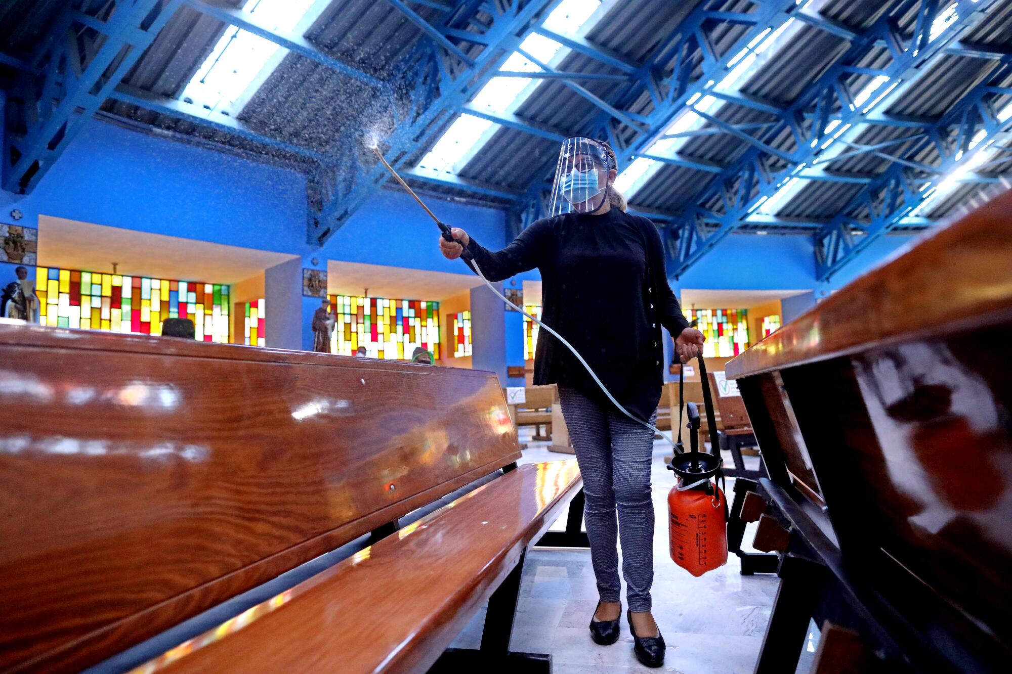 A woman in a mask and face shield uses a handheld pump to spray disinfectant on church pews