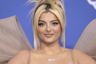 Bebe Rexha with a messy updo posing in a beige gown against a blue backdrop