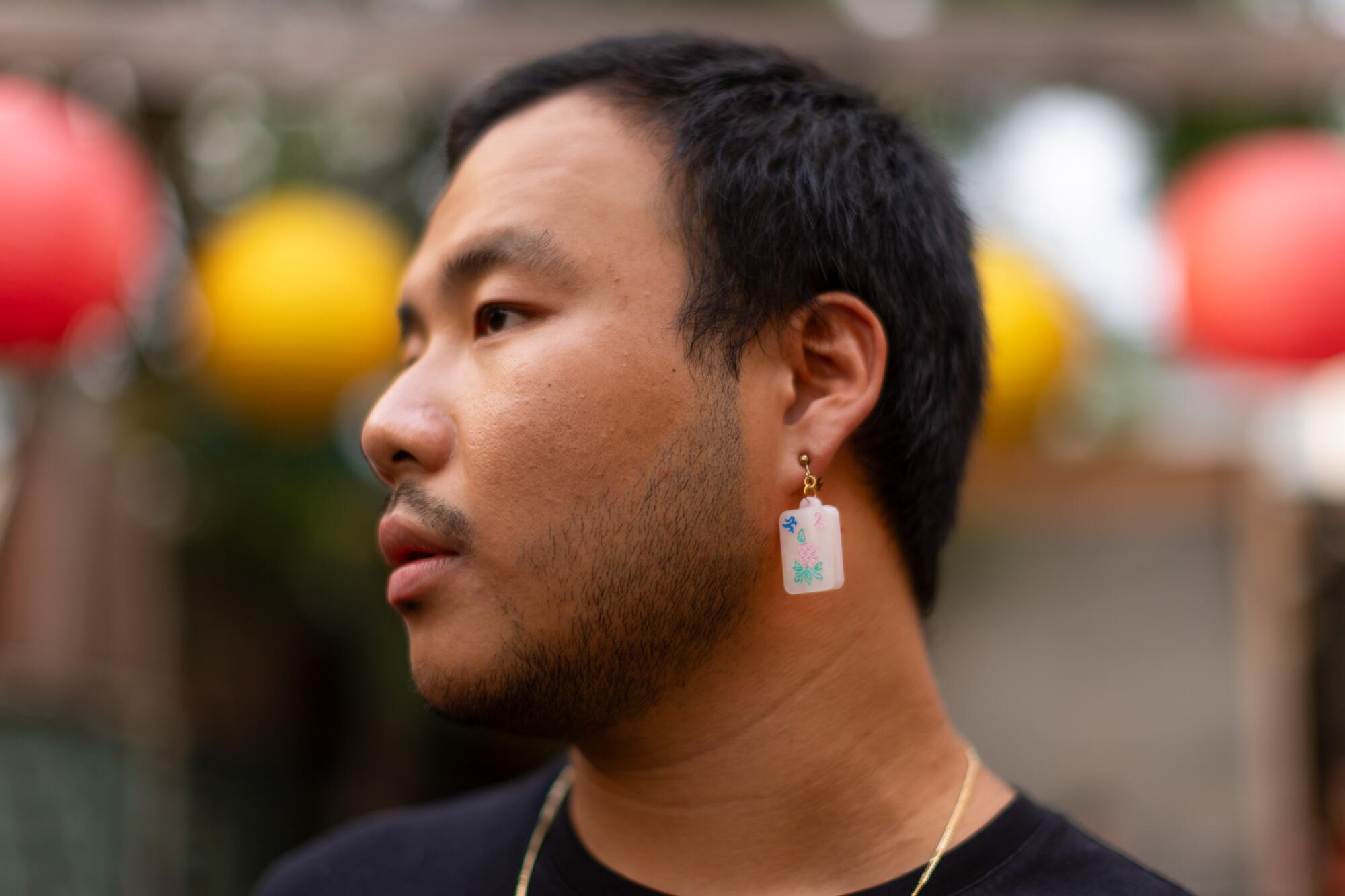 A person wears a mah-jongg-inspired earring that looks like a white tile  with blue, pink and green flower engravings.