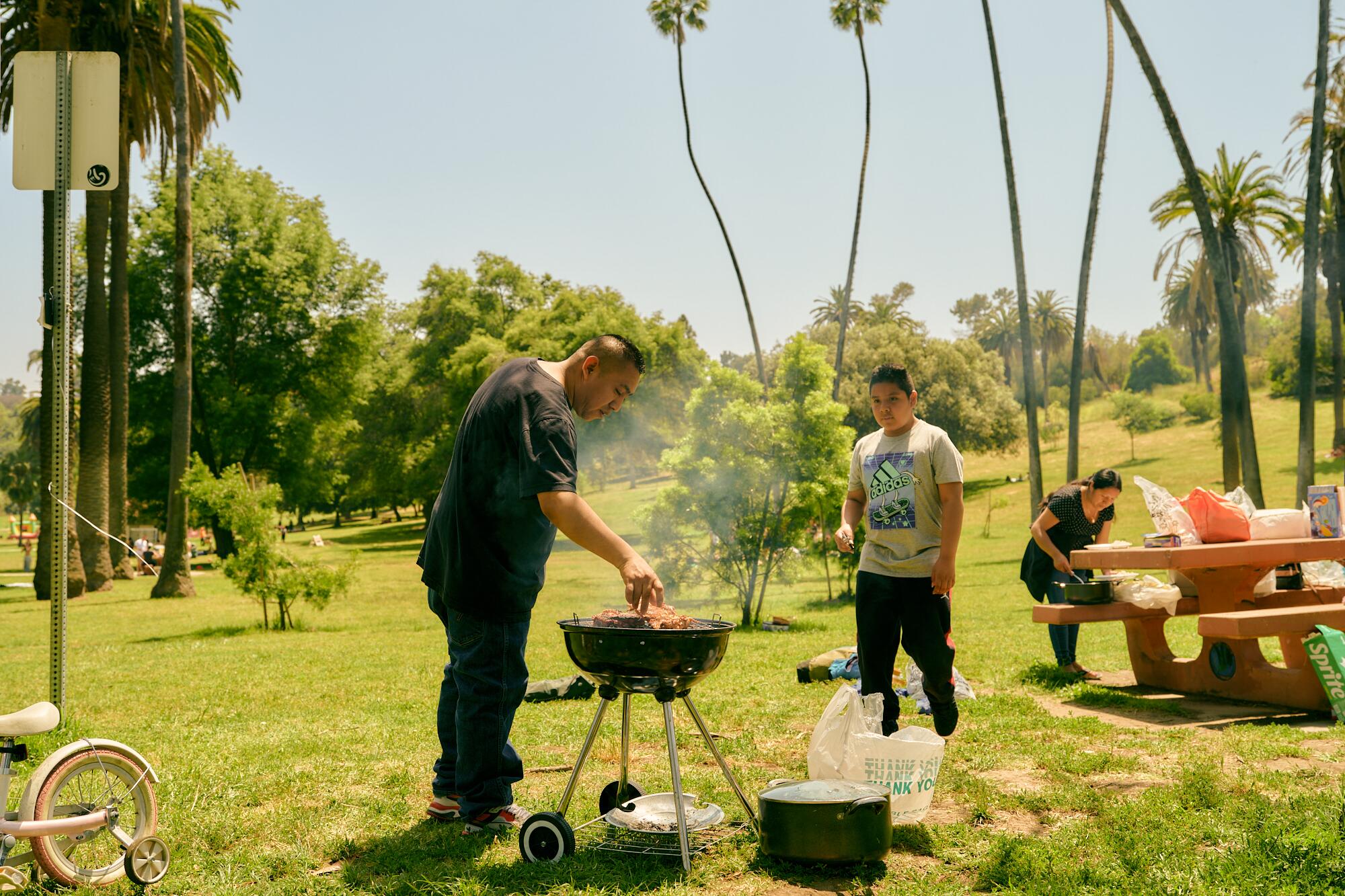 A man cooks meat on an outdoor grill next to a young boy and a woman at a picnic table.