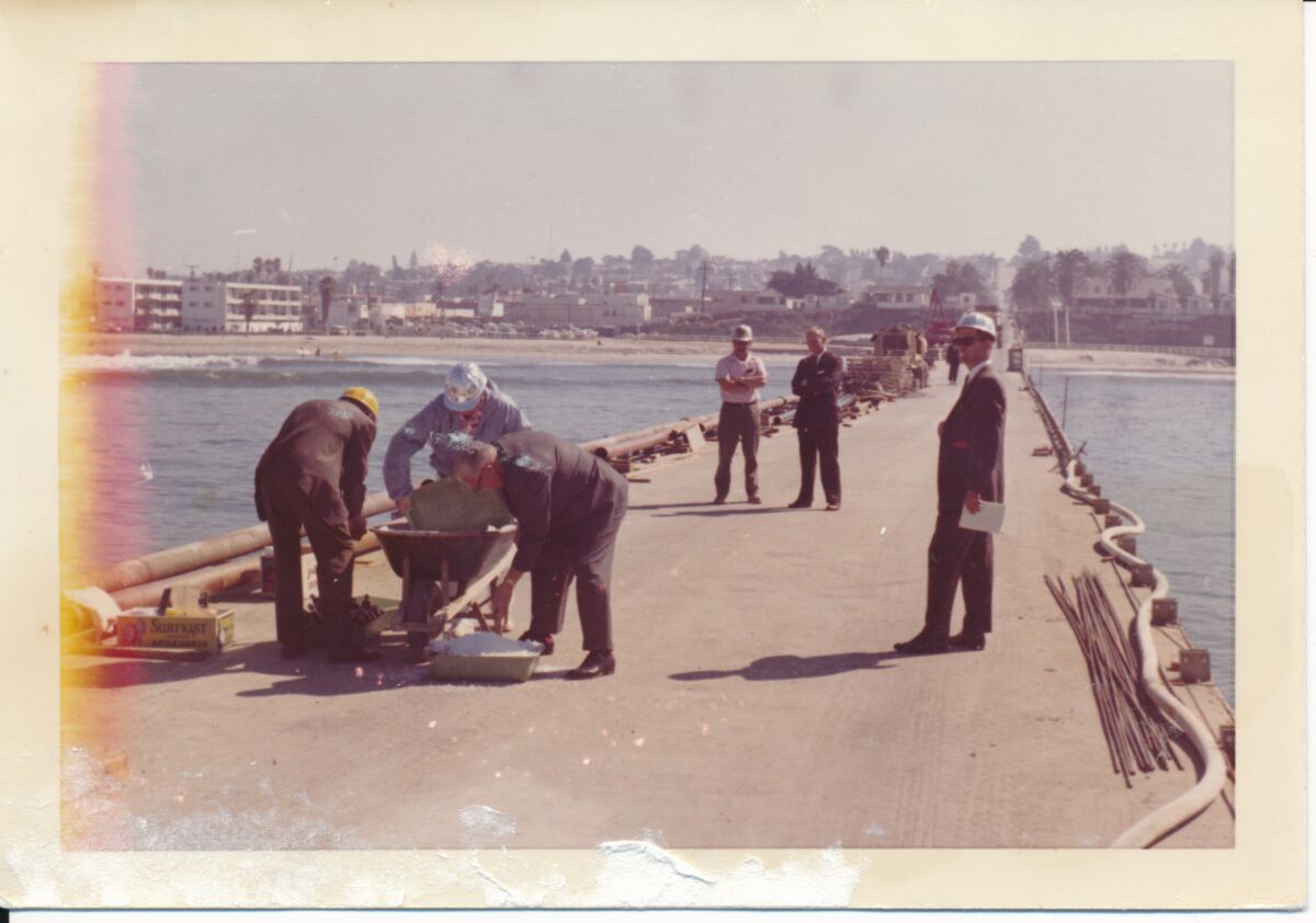 Faded archival photo of men in suits and hard hats on an ocean pier, beach in the background. Three stoop over a wheelbarrow.