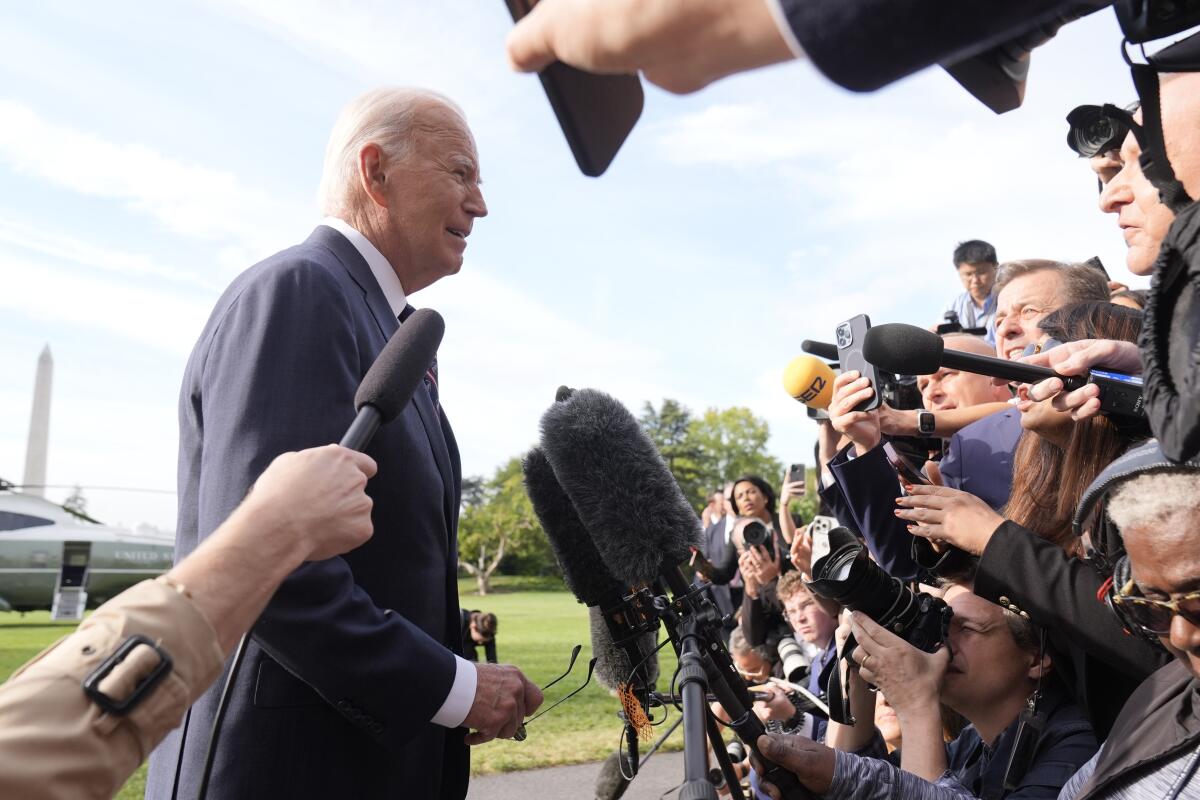 A gray-haired main, in dark suit and tie, seen from the side, speaks to a crowd of reporters