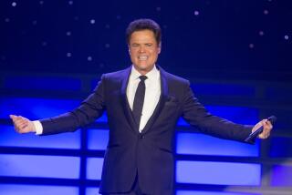 Donny Osmond on stage wearing a suit and tie smiling with his hands out to either side