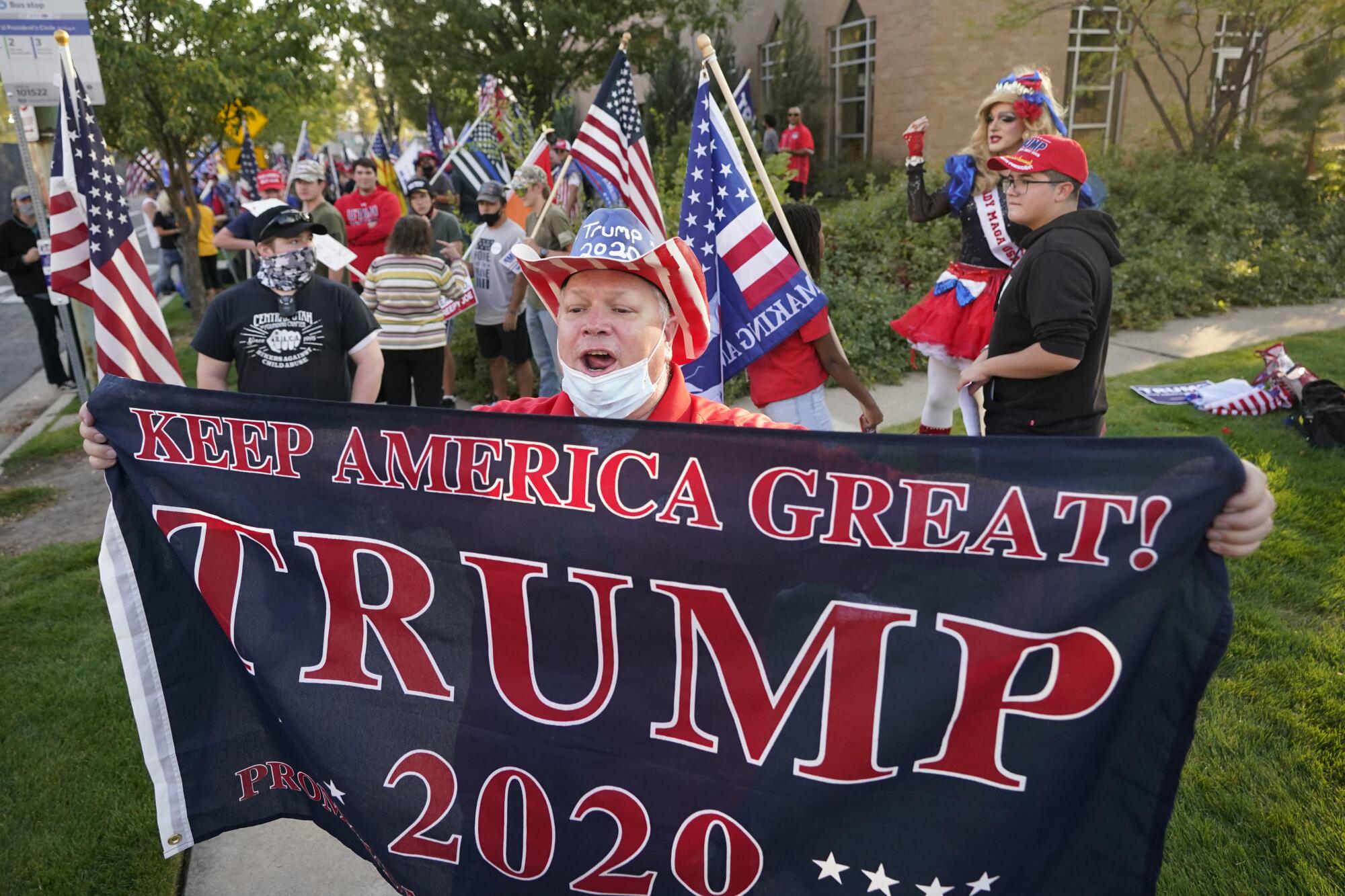 A person with a flag reading Keep America Great Trump 2020 in front of several people in U.S. flag gear