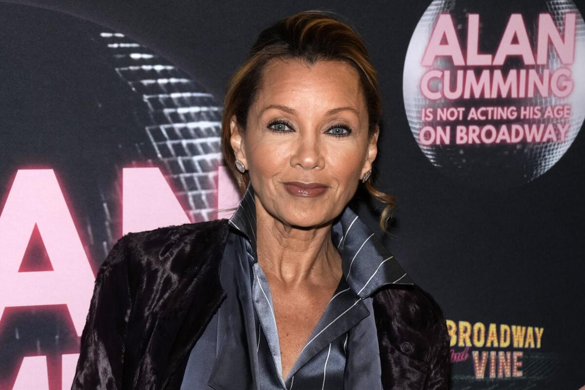 Vanessa Williams wearing a black velvet coat with a pinstriped gray shirt, standing in front of a dark backdrop