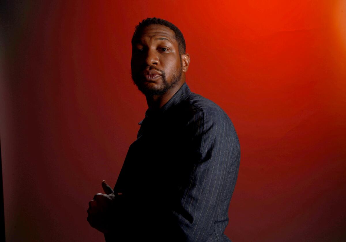 Jonathan Majors in a long-sleeve shirt posing in a side-profile against a dark red background