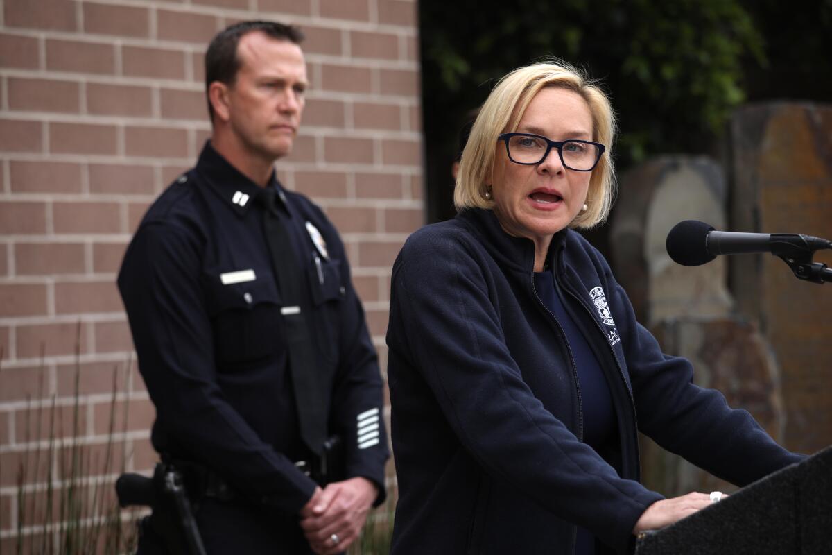 Traci Park, in dark jacket and black glasses, speaks at a podium. A man in police uniform stands nearby.