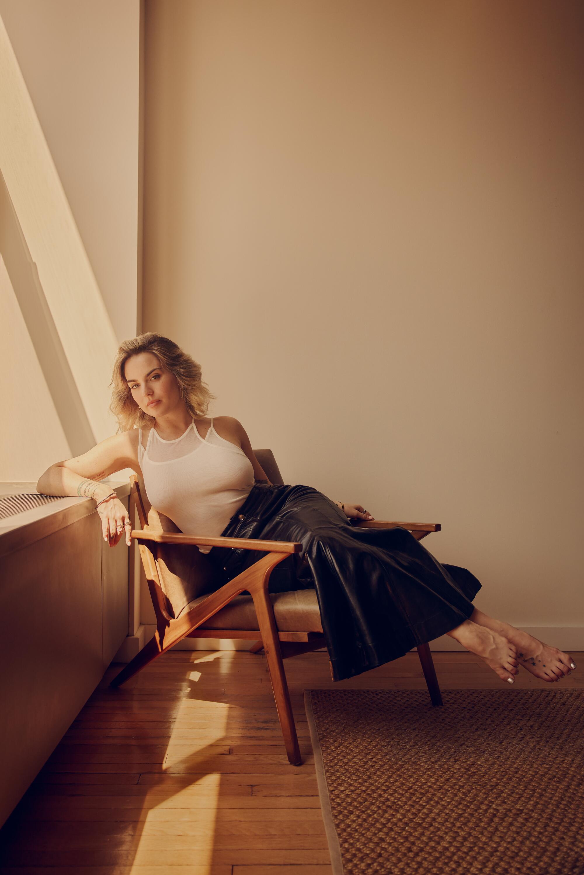 A blonde woman in a white T-shirt and a black leather skirt sits on a chair with her feet stretched out.