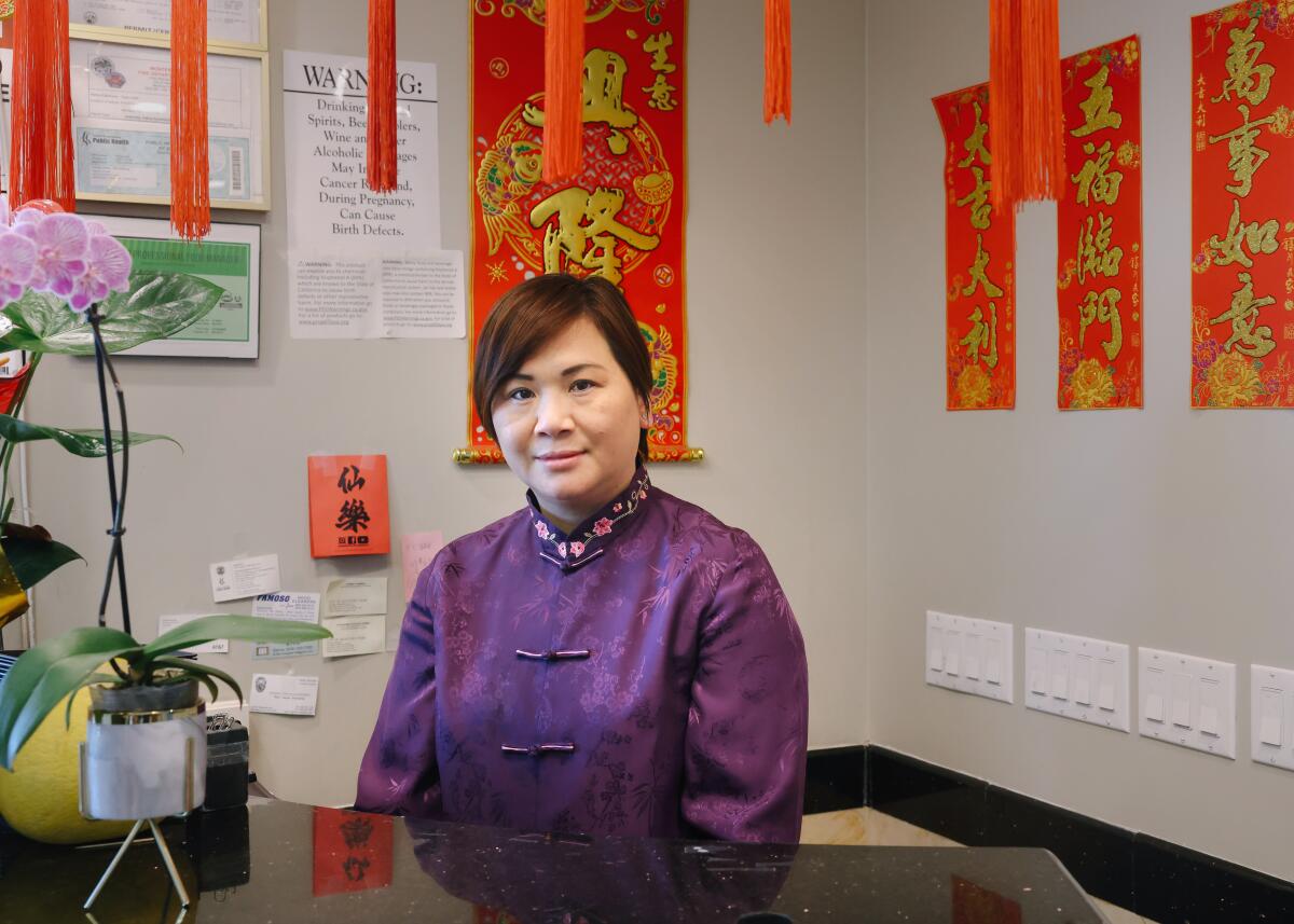 An Asian woman wearing purple silk shirt, lightly smiling sits behind a black marble counter, and red banners hang behind.