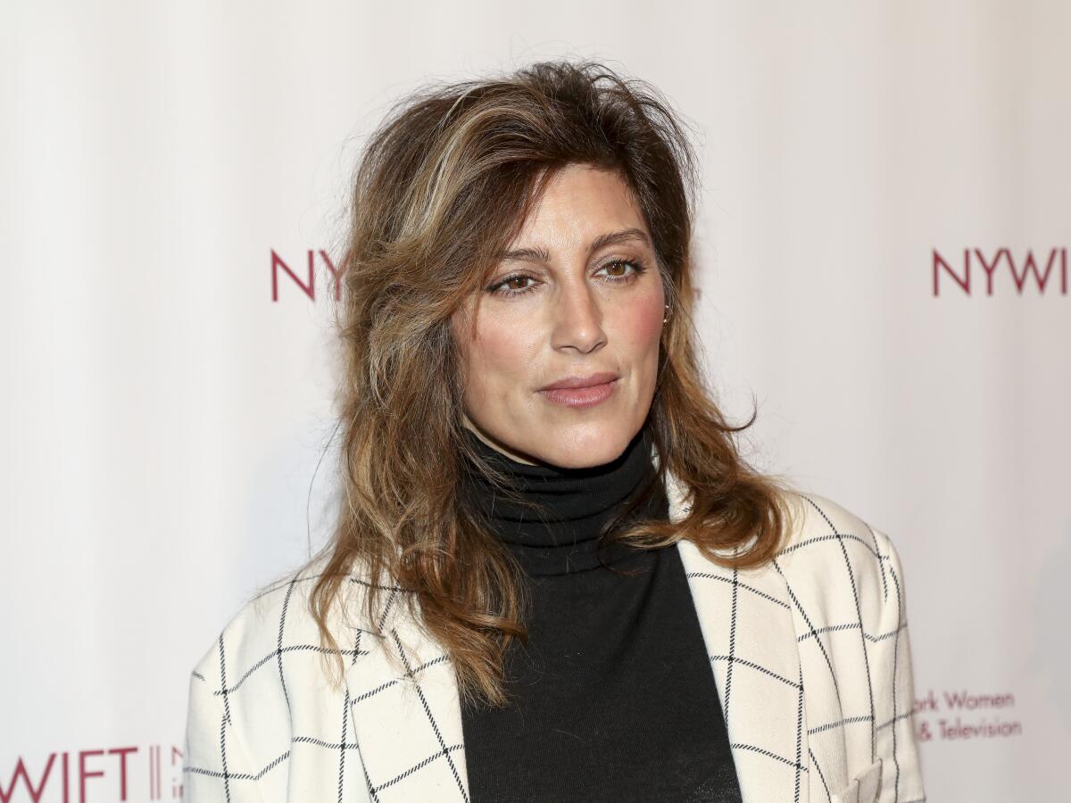 Jennifer Esposito looking serious while posing in a white plaid suit jacket over a black turtleneck
