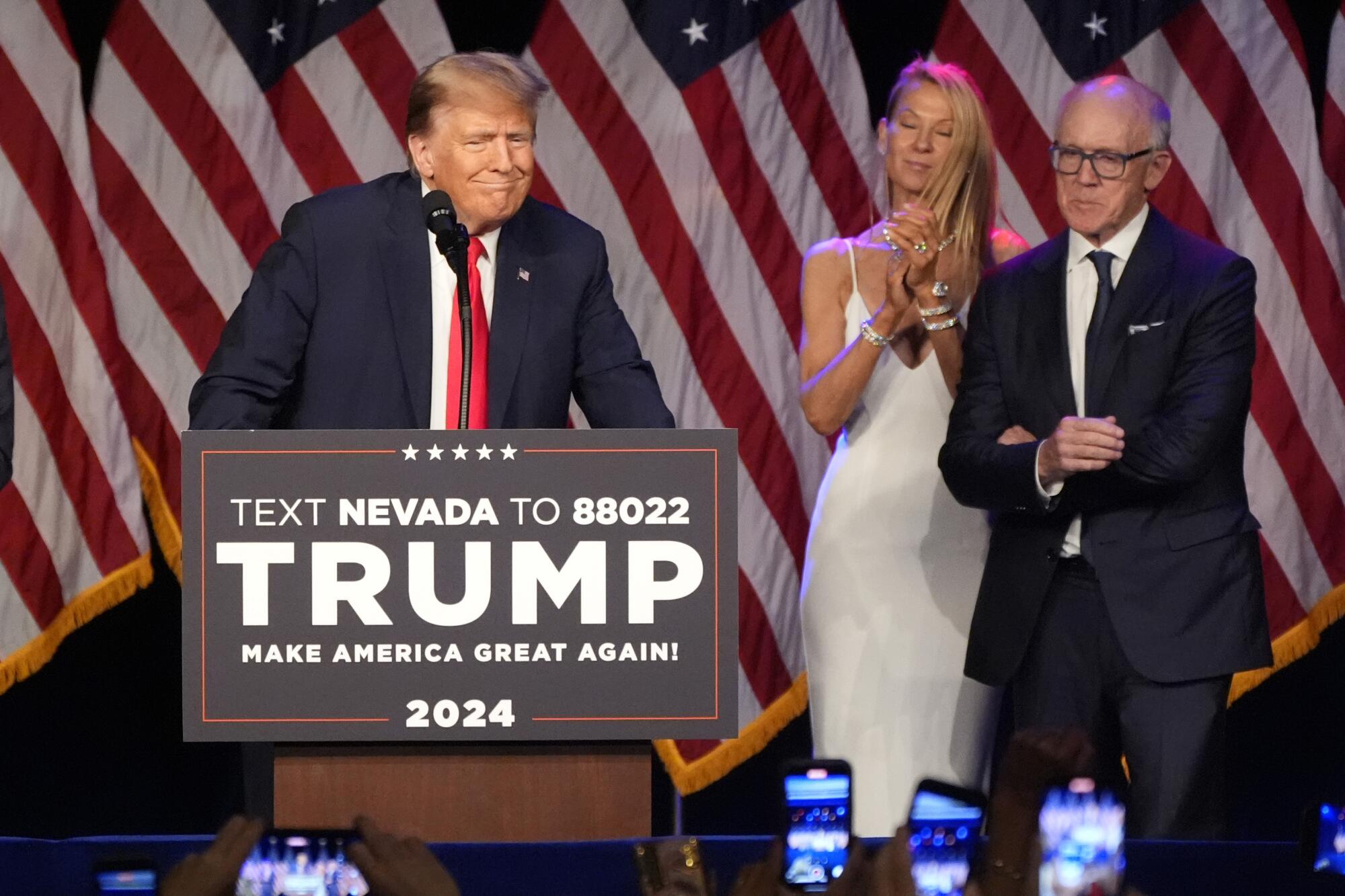 A man and woman stand by as Donald Trump speaks at a lectern with a campaign sign, in front of several draped American flags