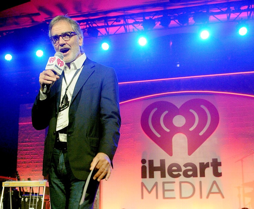 Bob Pittman, speaking into a microphone, stands on stage in front of a sign that says iHeartMedia.