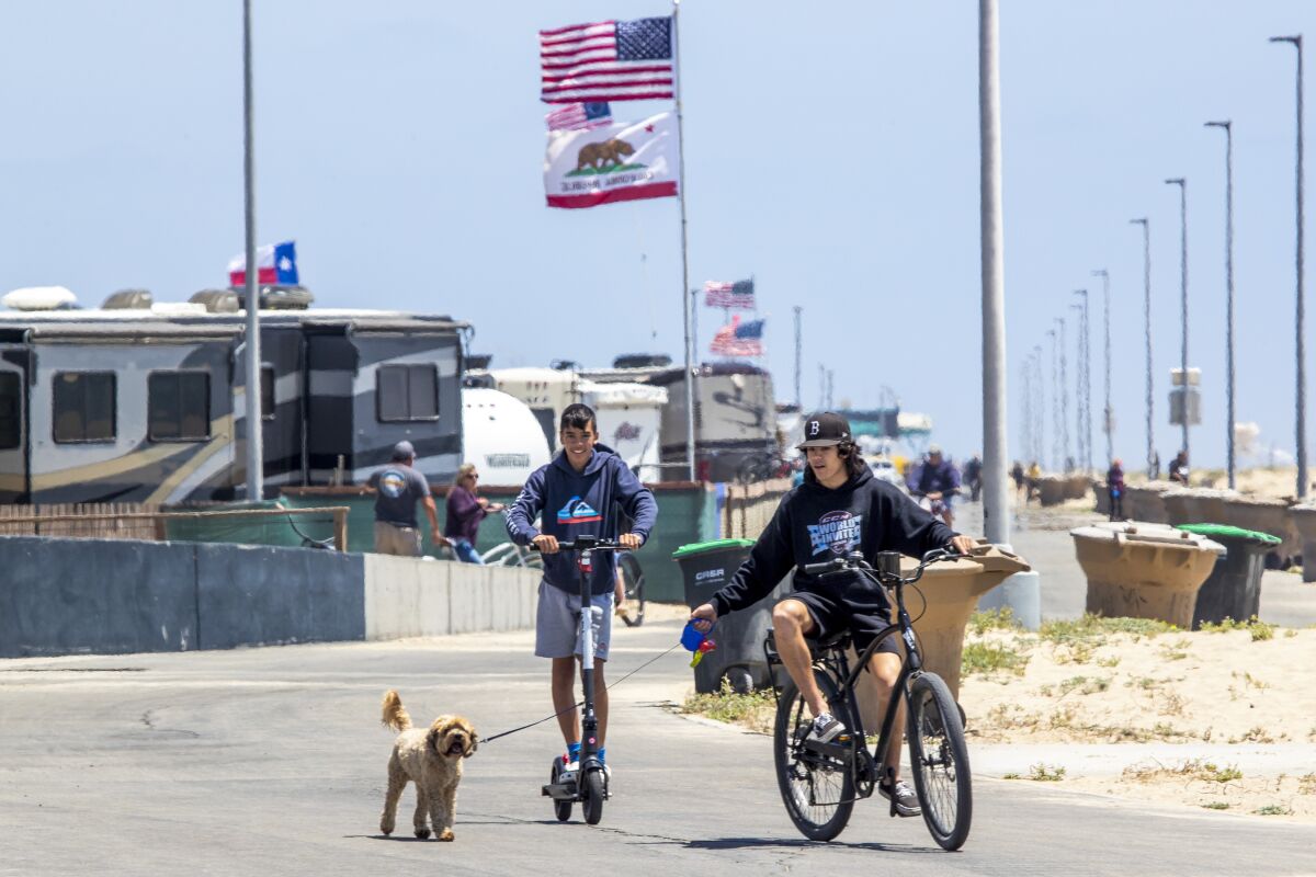 A person riding a beach cruiser bike with a dog on a leash, followed by a person on a scooter, on a beach path.
