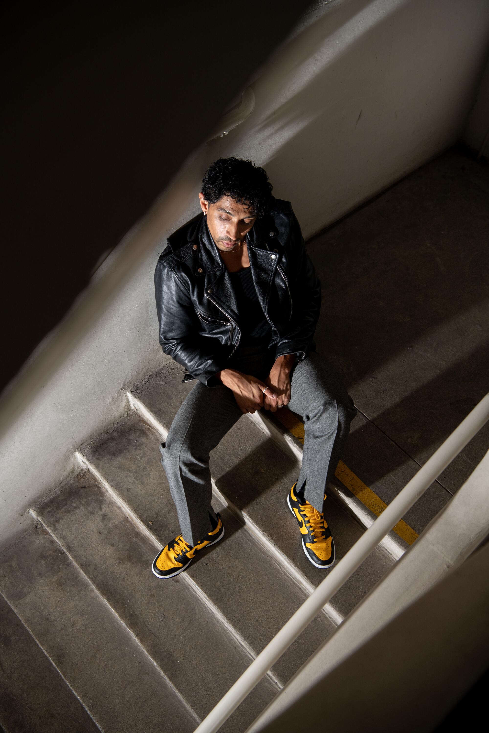 A man sits on stairwell wearing a leather jacket, gray pants, and yellow sneakers.