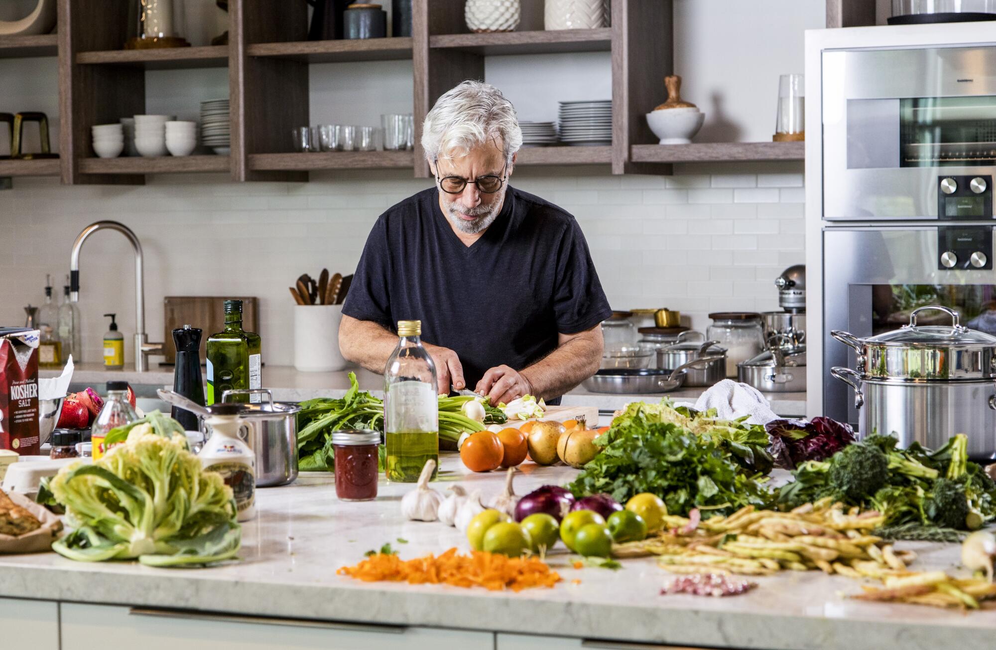 A man cuts vegetables on a kitchen island covered in vegetables.
