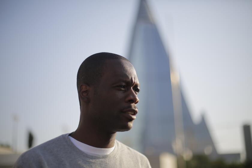 Pras Michel wearing a gray sweater and standing among city buildings