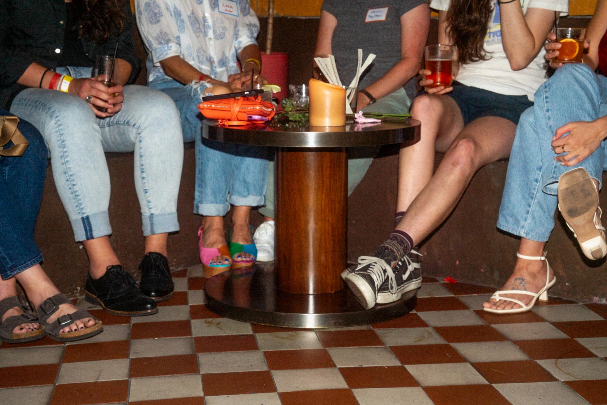Six sets of feet of people sitting around a small table full of cocktails and a candle