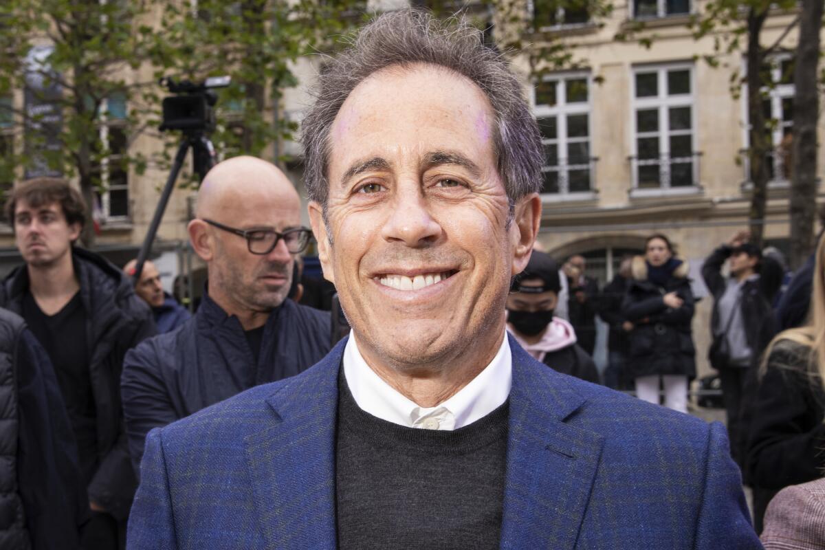 Jerry Seinfeld wears a blue blazer, grey sweater and a white collared shirt as he poses for a photo in front of a building