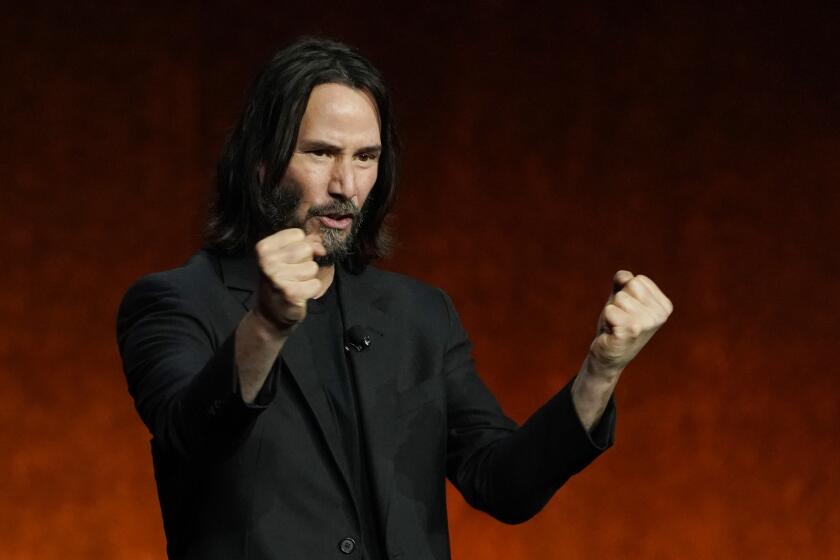 Man, Keanu Reeves, with long hair and bear, dressed in black blazer and shirt, clenching fists on stage