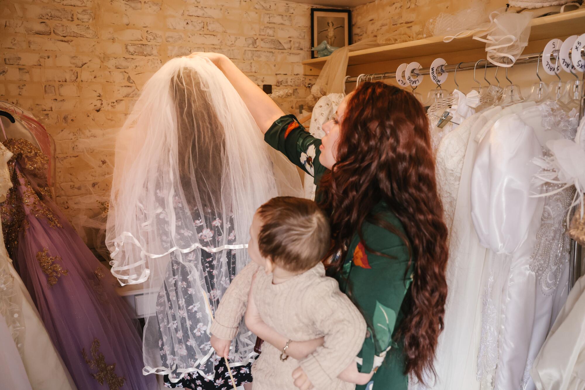 A woman holding a child helps another woman try on a veil