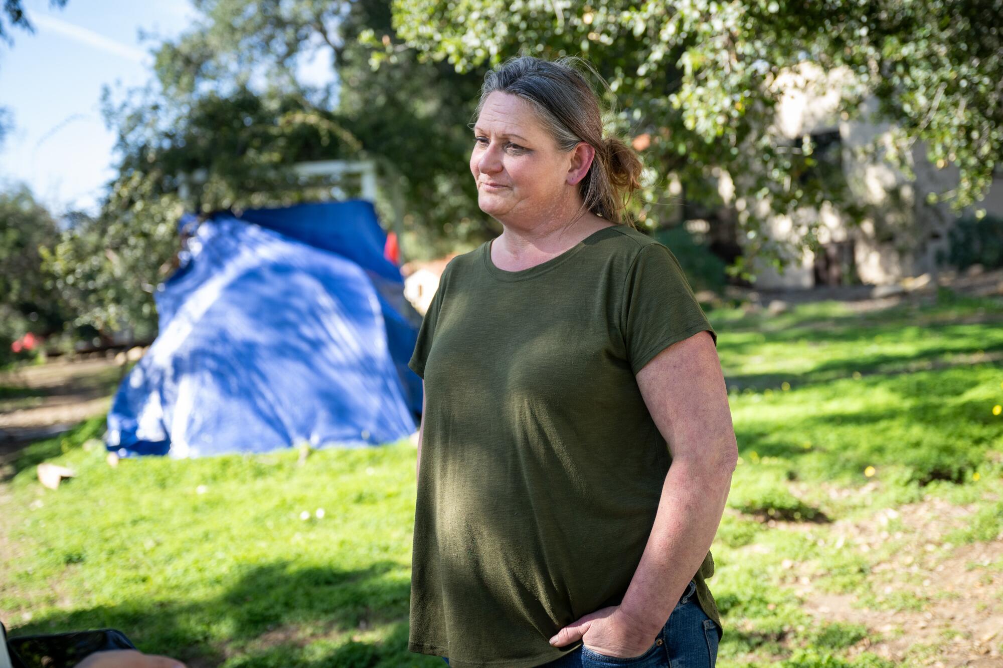 A woman stands near a blue tent on wooded property. 