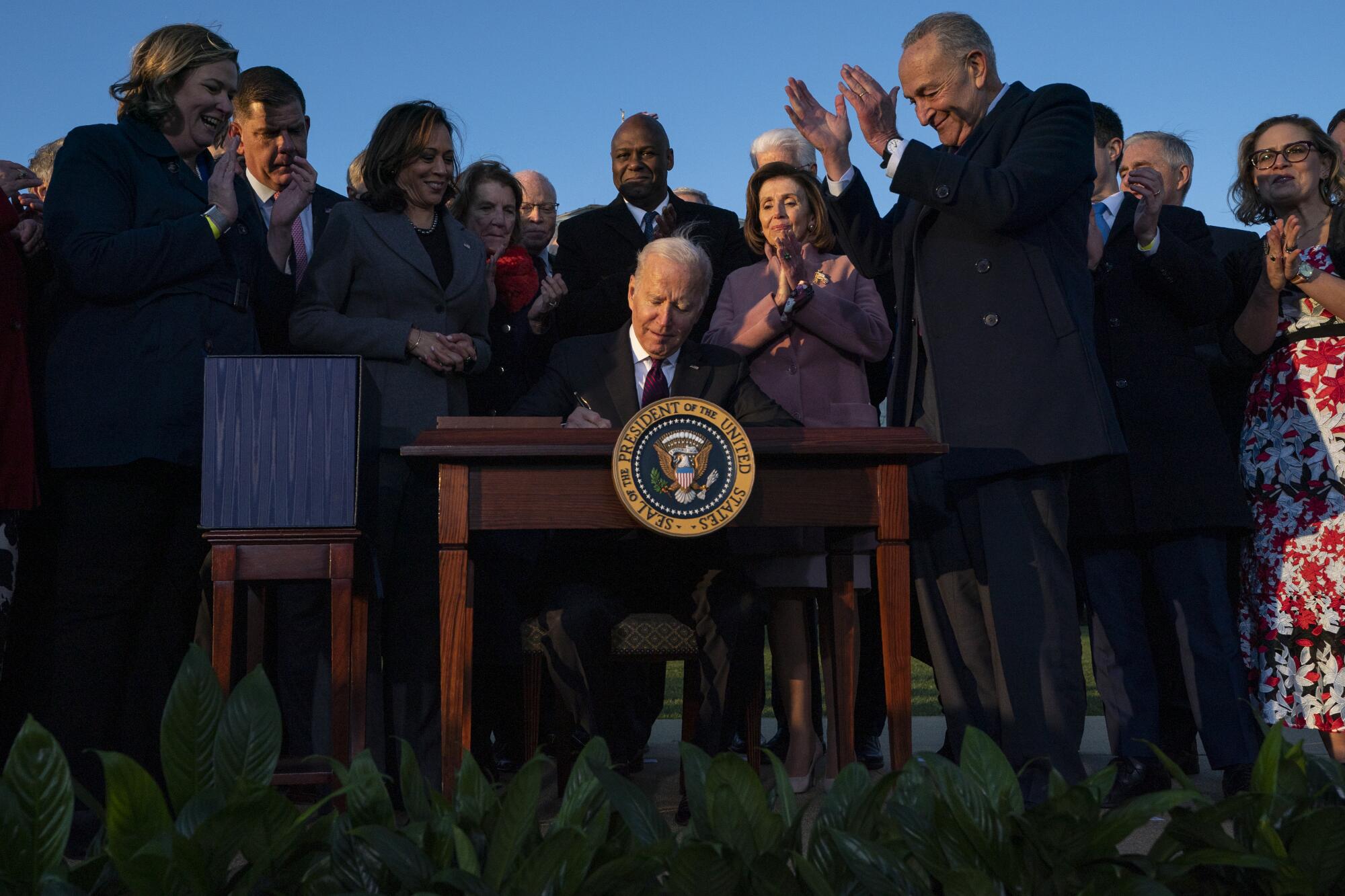 Lawmakers stand behind President Biden as he sits at desk and signs a bill