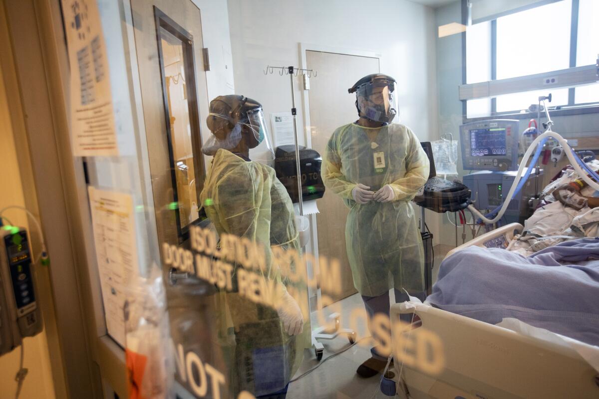 Two health workers in full protective gear observe a patient in an isolation room.