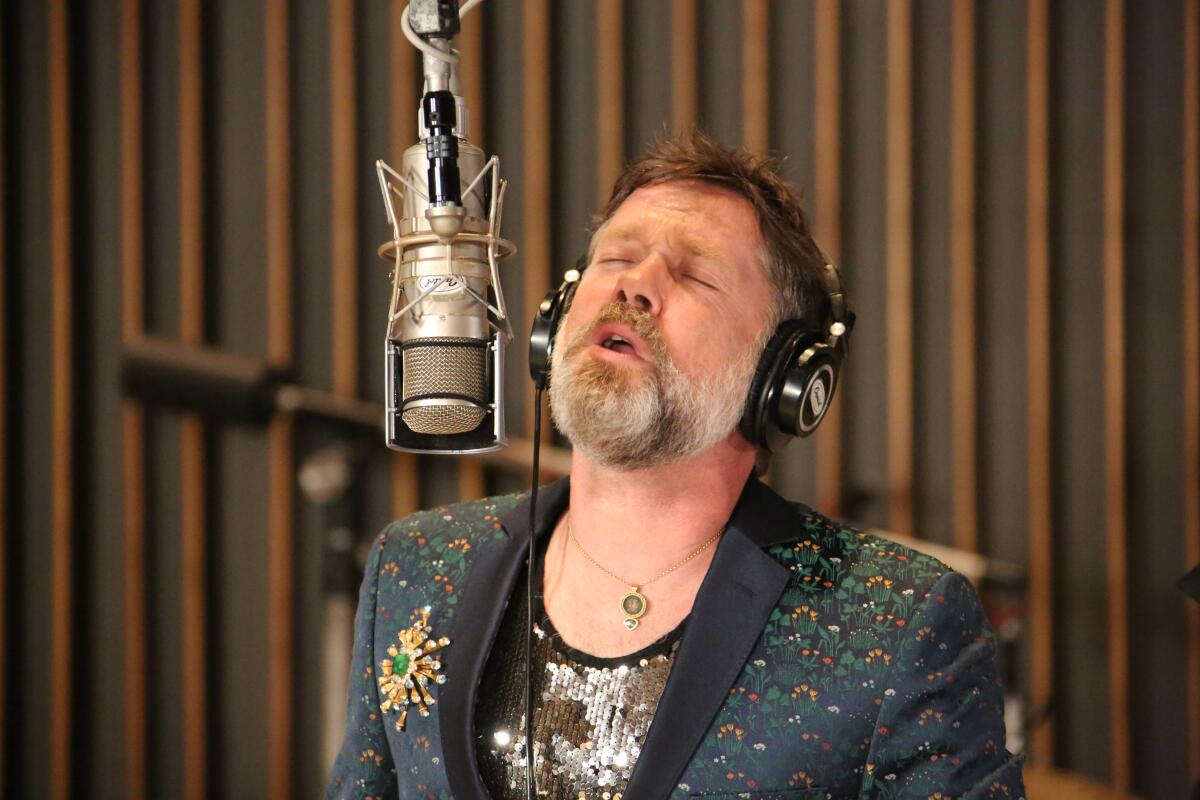A bearded man in a floral jacket and sequined shirt sings into a suspended microphone.