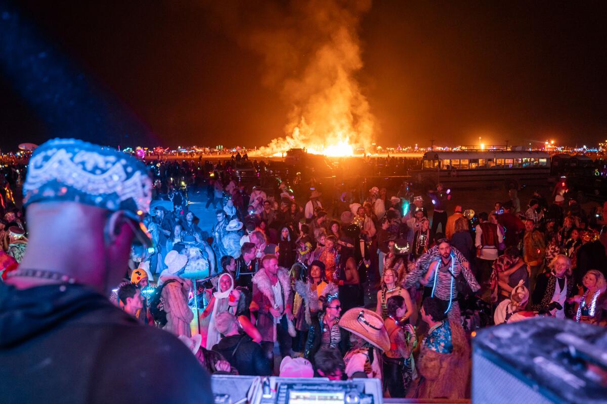 Que DJ plays music while several people dance in the dark and a giant fire gives off smoke in the background