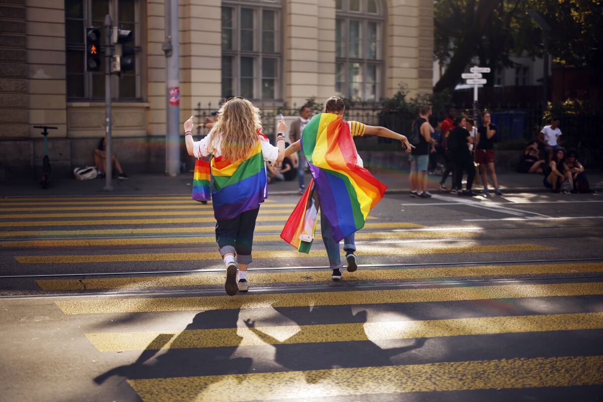 Two people draped in rainbow flags run across a street.