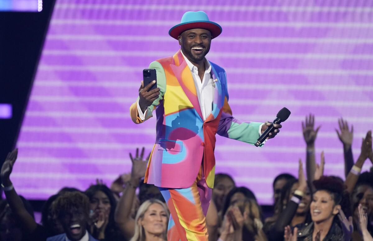 A smiling man in a multicolored suit and fedora holds a microphone and a cellphone as he walks on a stage.