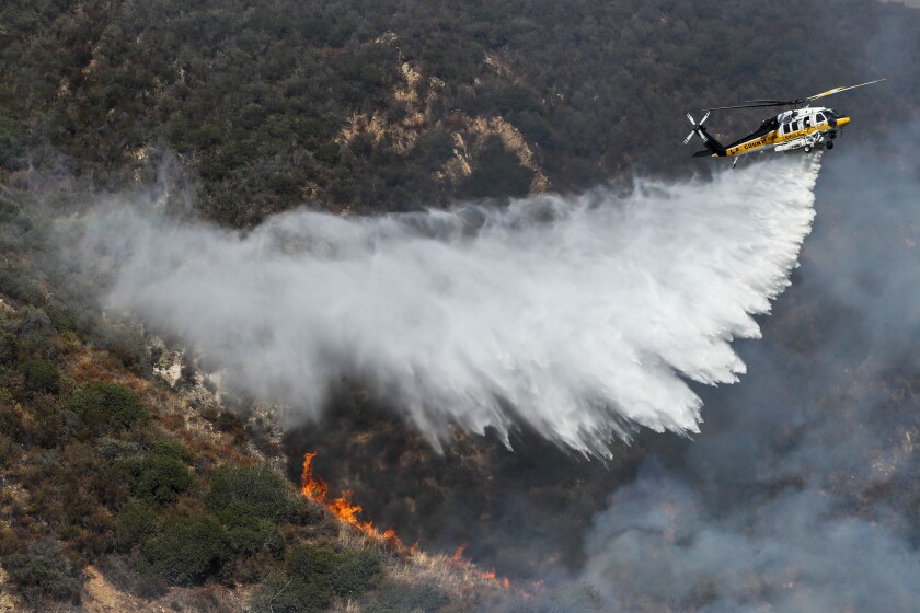 A helicopter throws water over a line of flames on a hill