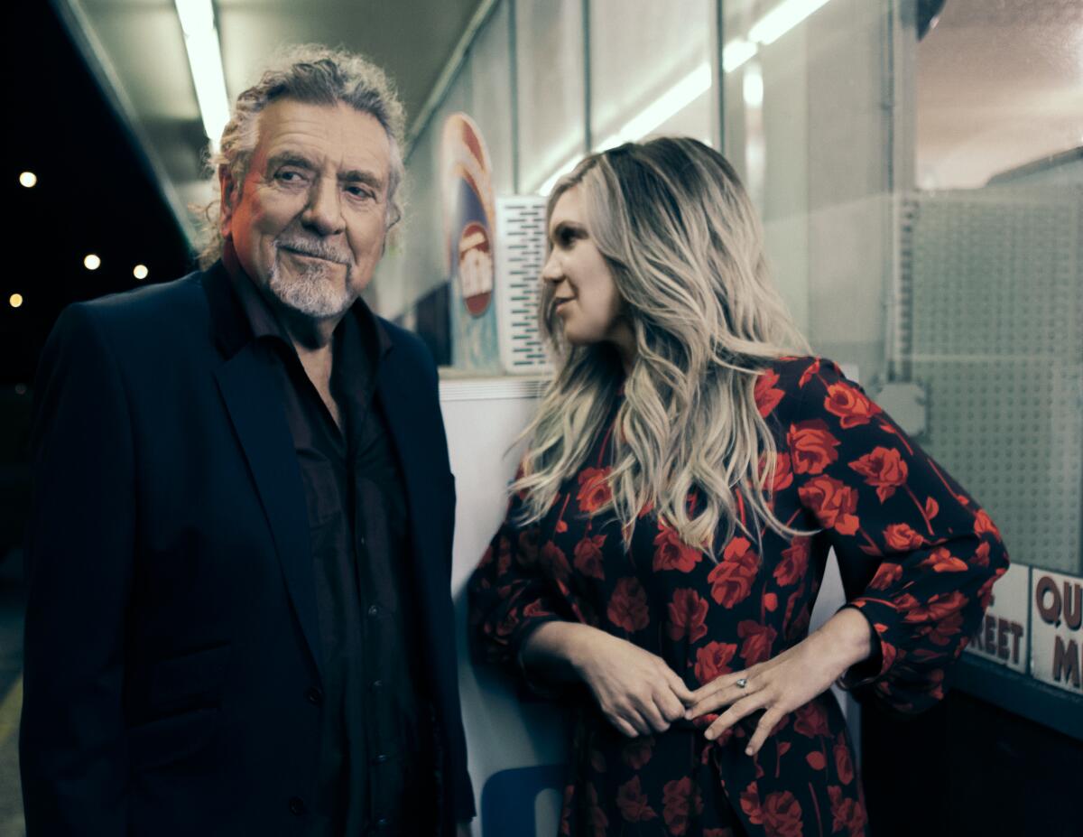 An older man in a dark sports coat and shirt stands next to a blond woman in a flowered dress