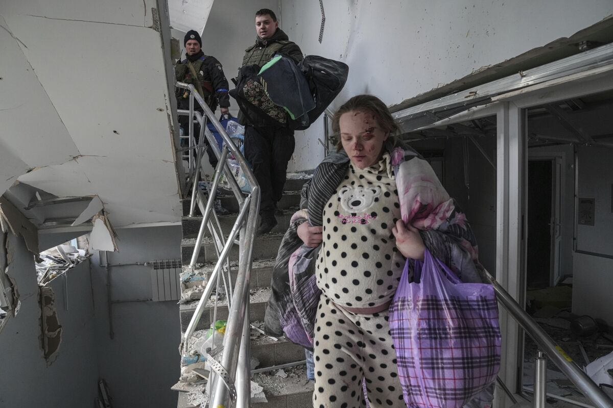 A pregnant woman with scrapes on her face walks down damaged stairs in her pajamas, carrying her belongings
