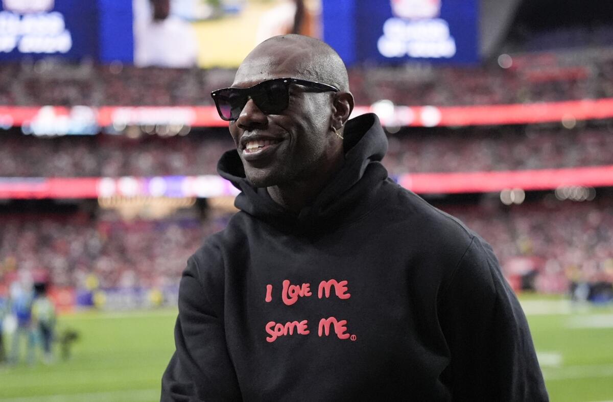 Terrell Owens smiles while wearing shades and a hoodie that reads "I Love Me Some Me" and standing on the field before a game