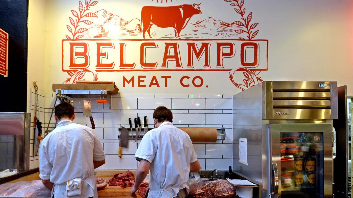 Two men work at a wood counter covered with raw meat under a "Belcampo Meat Co." sign