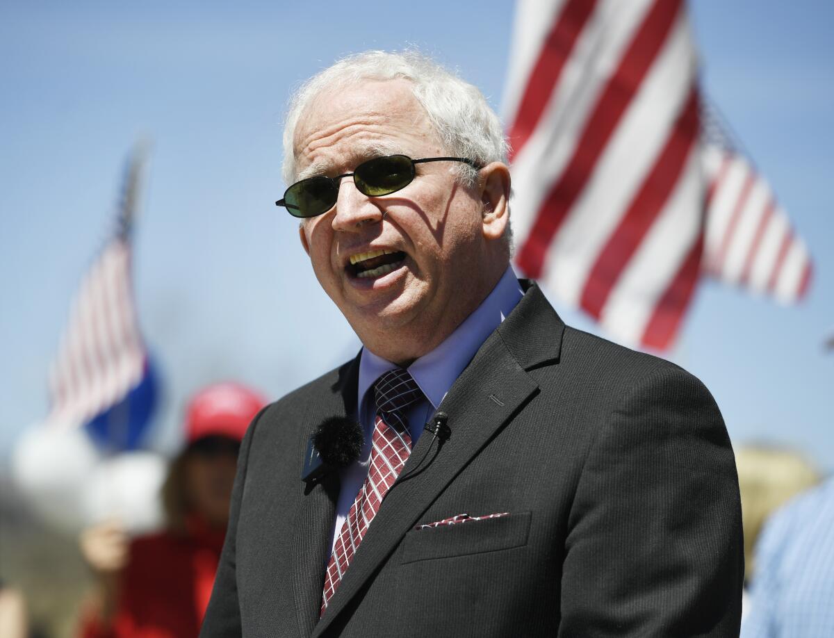 A man talks outdoors with American flags behind him.