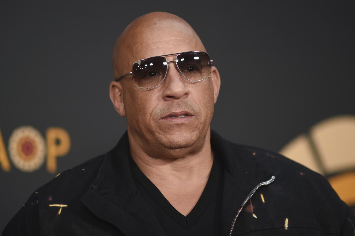 Vin Diesel wears dark sunglasses and a black formal shirt and poses with a straight face against a dark background