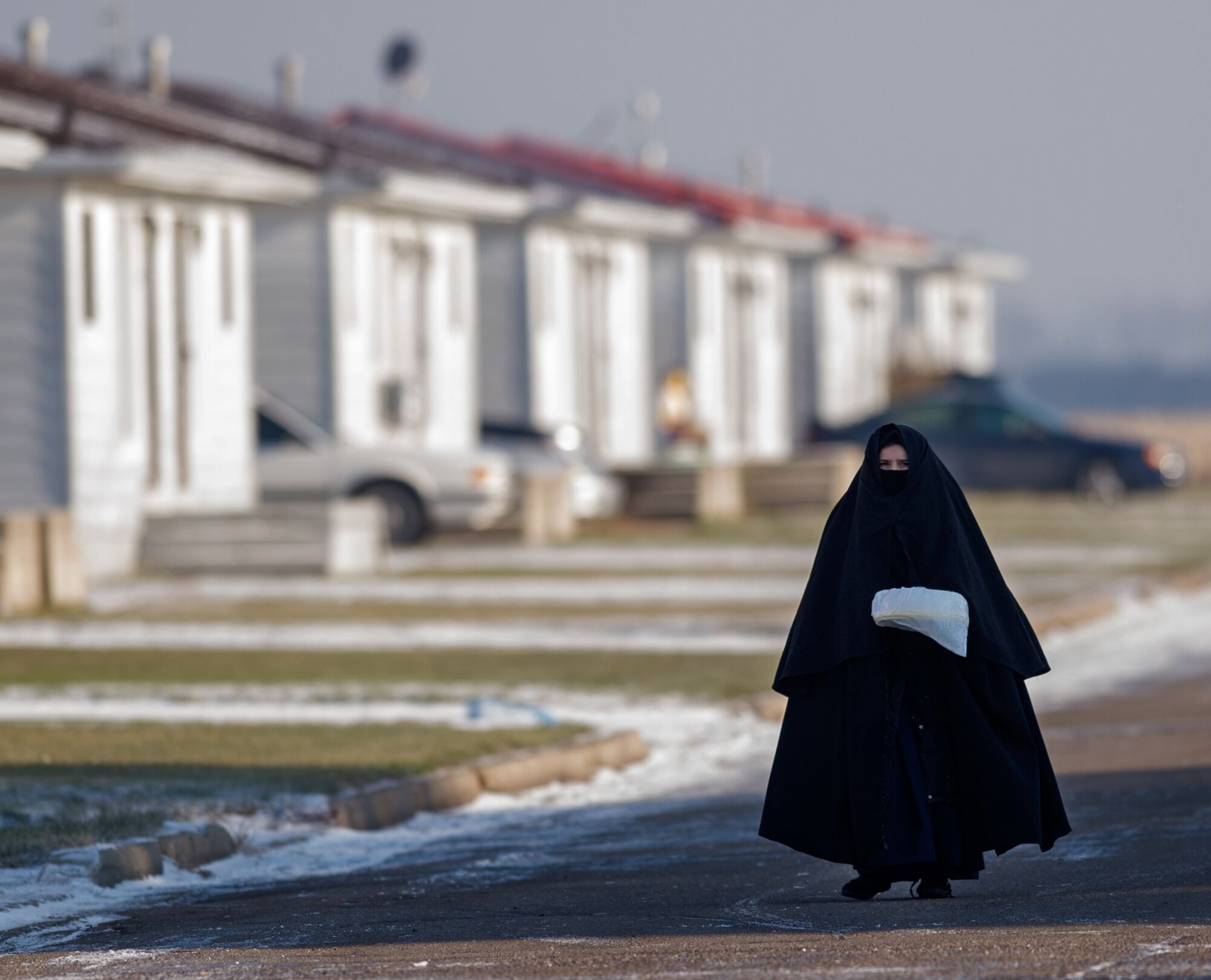 A woman wearing a flowing black robe and head covering walks past homes on the left, with snow on the ground